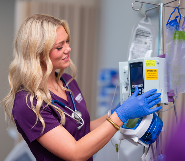 Student in Mercy Health Sciences University scrubs adjusting settings on an intravenous infusion pump while practicing patient care skills in a simulated clinical environment.