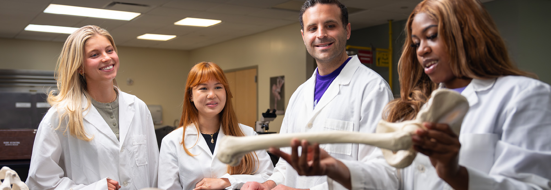 Group of students in laboratory coats gathered around while studying a large anatomical bone model in a health sciences lab classroom.
