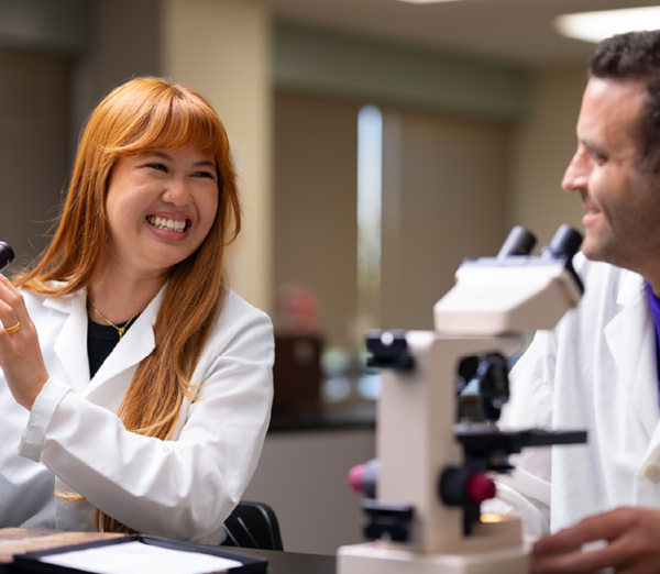 Students in laboratory coats working with a microscope and discussing observations during a health sciences lab class.