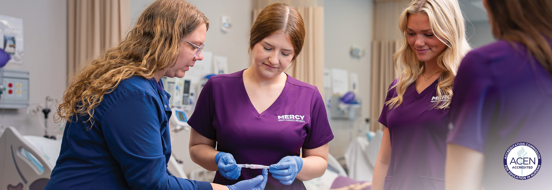Faculty member demonstrating a clinical skill to nursing students wearing Mercy Health Sciences University scrubs and examination gloves in a simulated patient care environment.