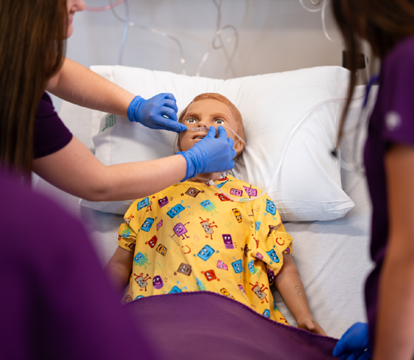 Students in clinical training adjusting oxygen tubing on a patient simulation manikin positioned in a hospital bed during a nursing skills exercise.