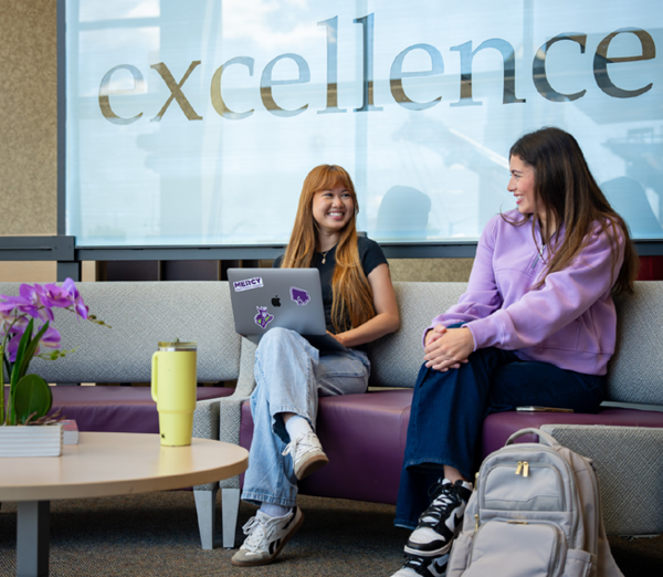Two people seated in the Sullivan Center lobby lounge area, talking beside an open laptop, with seating, tables, and a large “excellence” graphic in the background.