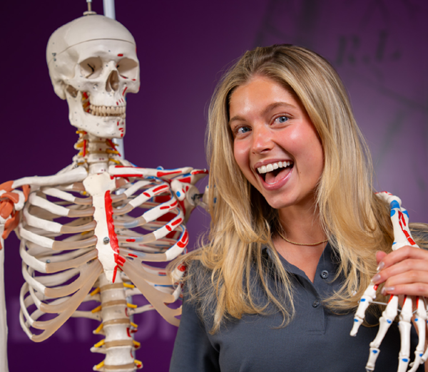 Person posing beside an anatomical skeleton model, with the model’s arm positioned around the person’s shoulder in a classroom setting.