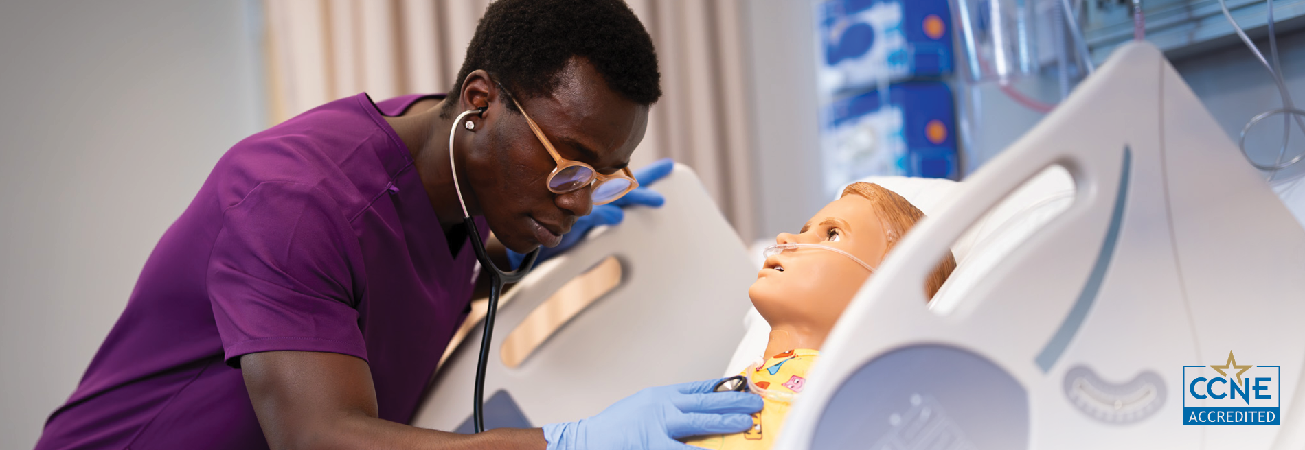 Student in Mercy Health Sciences University scrubs and gloves using a stethoscope to examine a patient simulation manikin in a healthcare simulation lab.