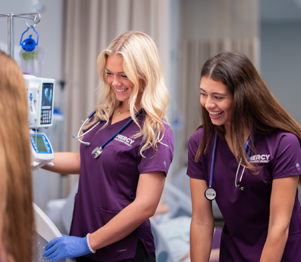 Students in Mercy Health Sciences University scrubs working together at a patient bedside simulation station, with monitoring equipment and clinical supplies visible.