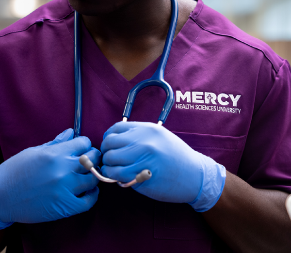 Close-up of a healthcare student in Mercy Health Sciences University scrubs wearing examination gloves and holding a stethoscope.