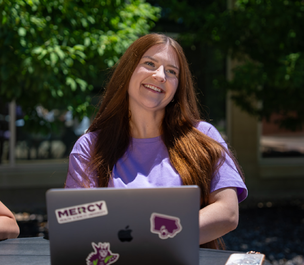 Person seated outdoors at a table using a Mercy-branded laptop, with trees and campus landscaping in the background.