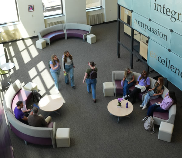 Overhead view of students walking and meeting in the Sullivan Center lobby, featuring collaborative seating areas and a wall displaying the words “compassion” and “excellence.”