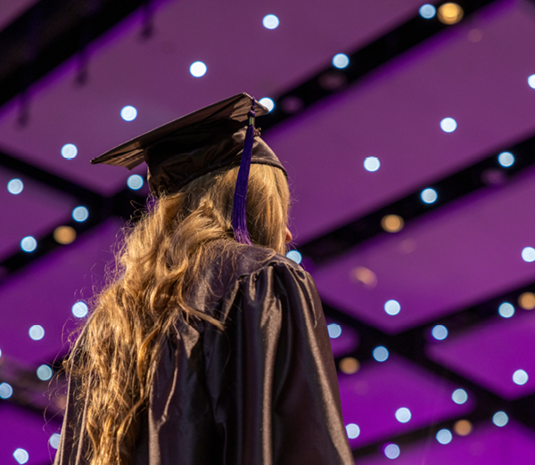 Graduate in cap and gown viewed from behind during commencement, standing beneath purple stage lighting and ceiling lights in the ceremony venue.