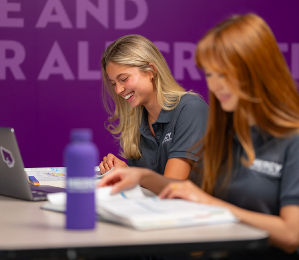 Students reviewing course materials at a table with open textbooks, a laptop, and a water bottle, seated in front of a purple wall.