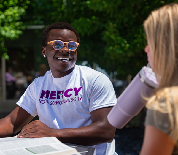 Student studying outdoors with an open textbook, talking with a classmate at a campus table.