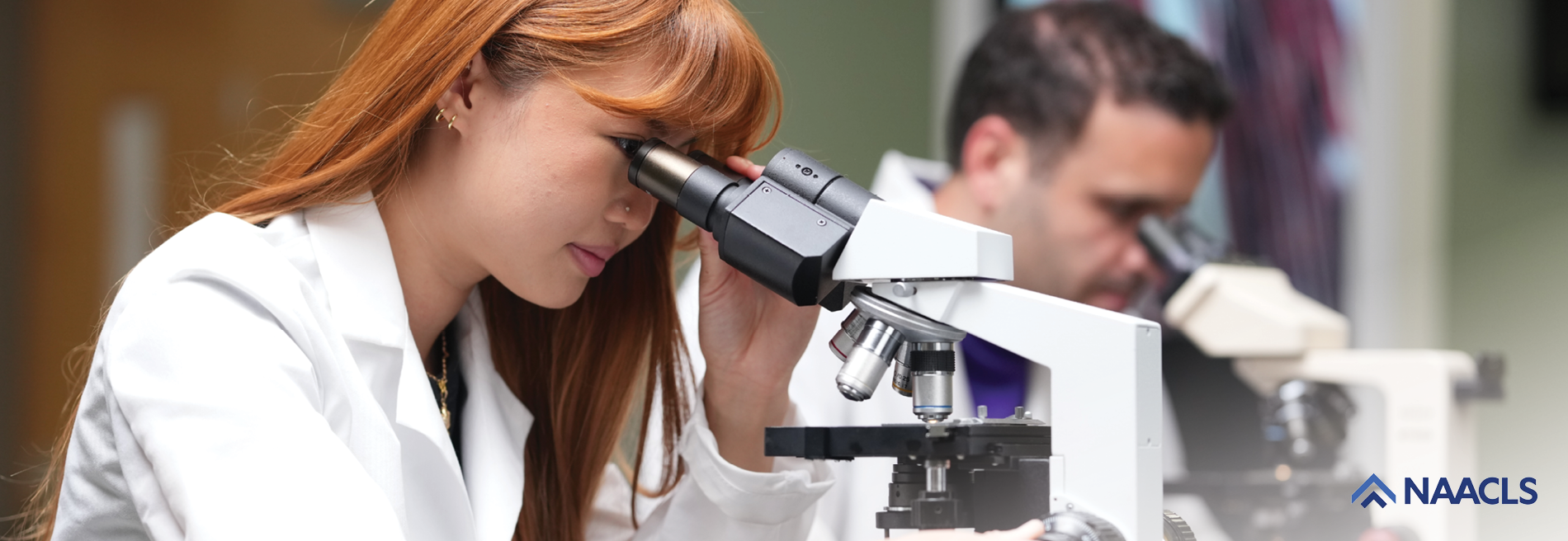 Close-up of a student in a laboratory coat examining a specimen through a microscope while another student works at a nearby microscope station.