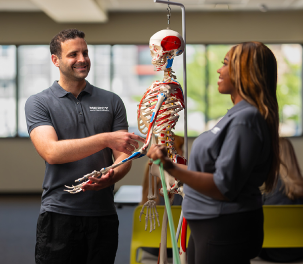 Students participating in a physical therapist assistant training session with an anatomical skeleton model and exercise bands in a healthcare learning environment.