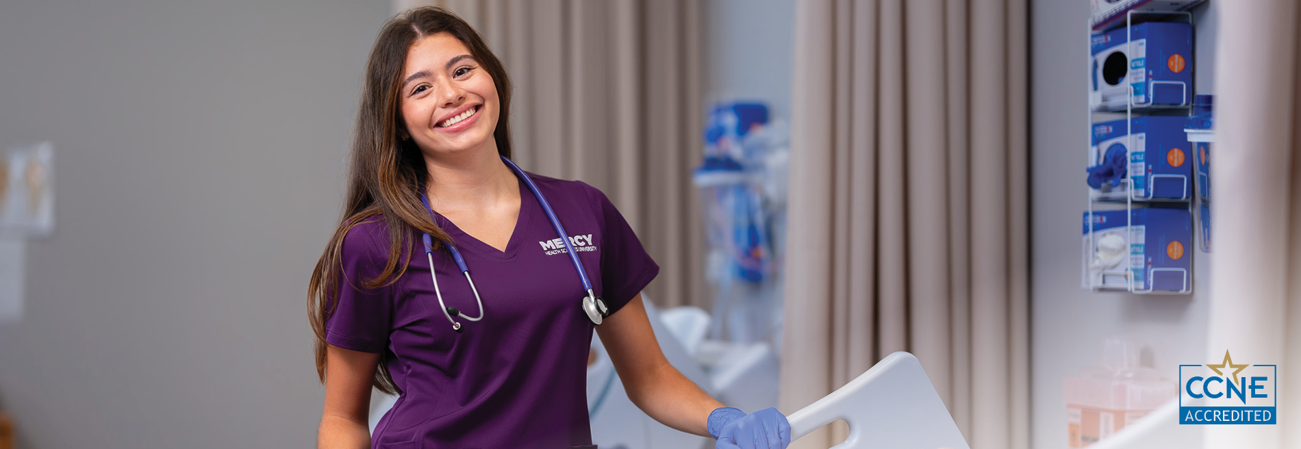 Student in purple Mercy Health Sciences University scrubs and a stethoscope standing in a healthcare simulation environment, with a hospital bed and clinical supplies visible in the background.