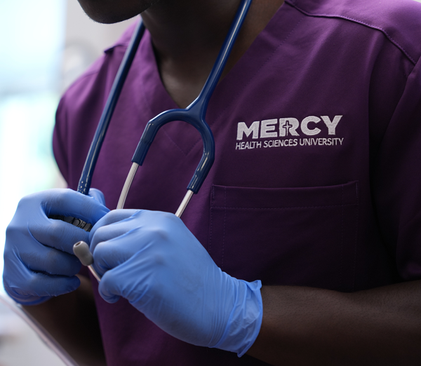 Nursing student in purple Mercy Health Sciences University scrubs adjusting a stethoscope while wearing examination gloves.