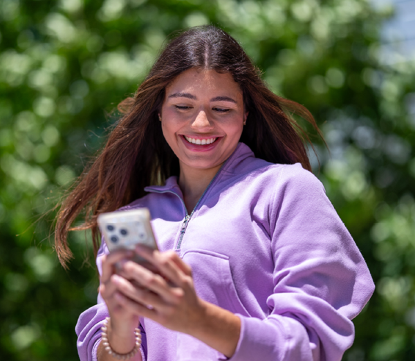Student outdoors on campus looking at a smartphone, with trees and greenery in the background.