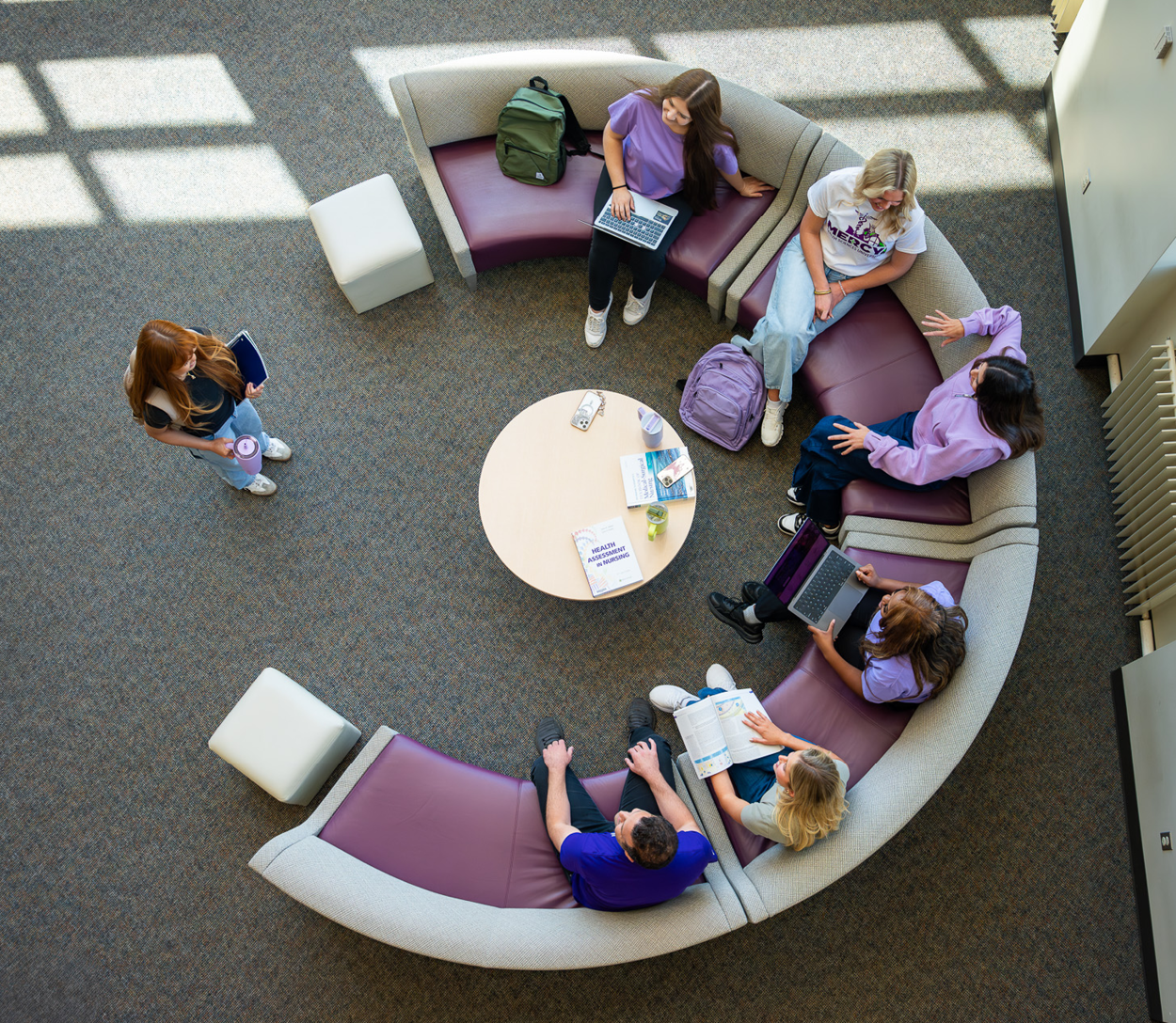 Overhead view of several Mercy Health Sciences University students seated in a circular lounge area, studying, using laptops, reading textbooks, and collaborating around a central table in a bright campus common space.
